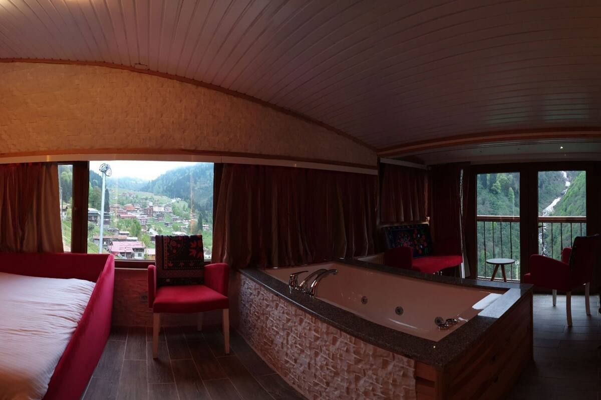 A bedroom with a large bed, a red chair, a bathtub, and a view of a village through large windows.