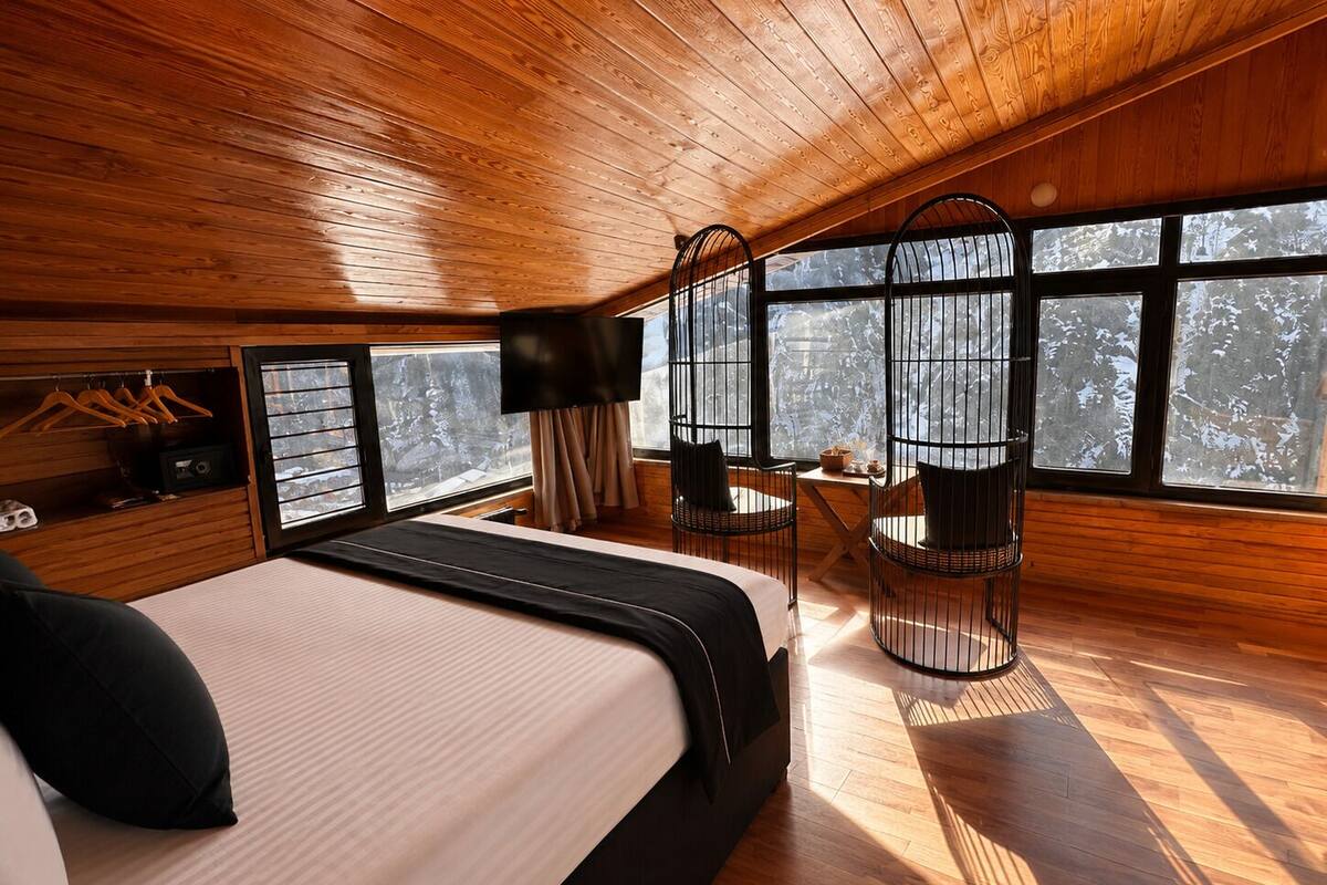 A wooden cabin bedroom with a bed, two chairs, a table, a TV, and large windows.