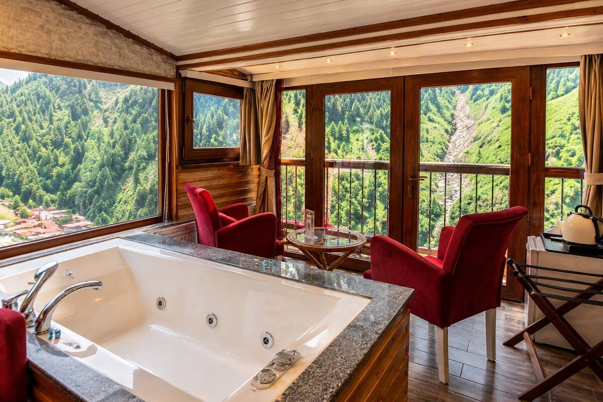 A room with a large bathtub, red chairs, a round table, and a view of a forested landscape through large windows.