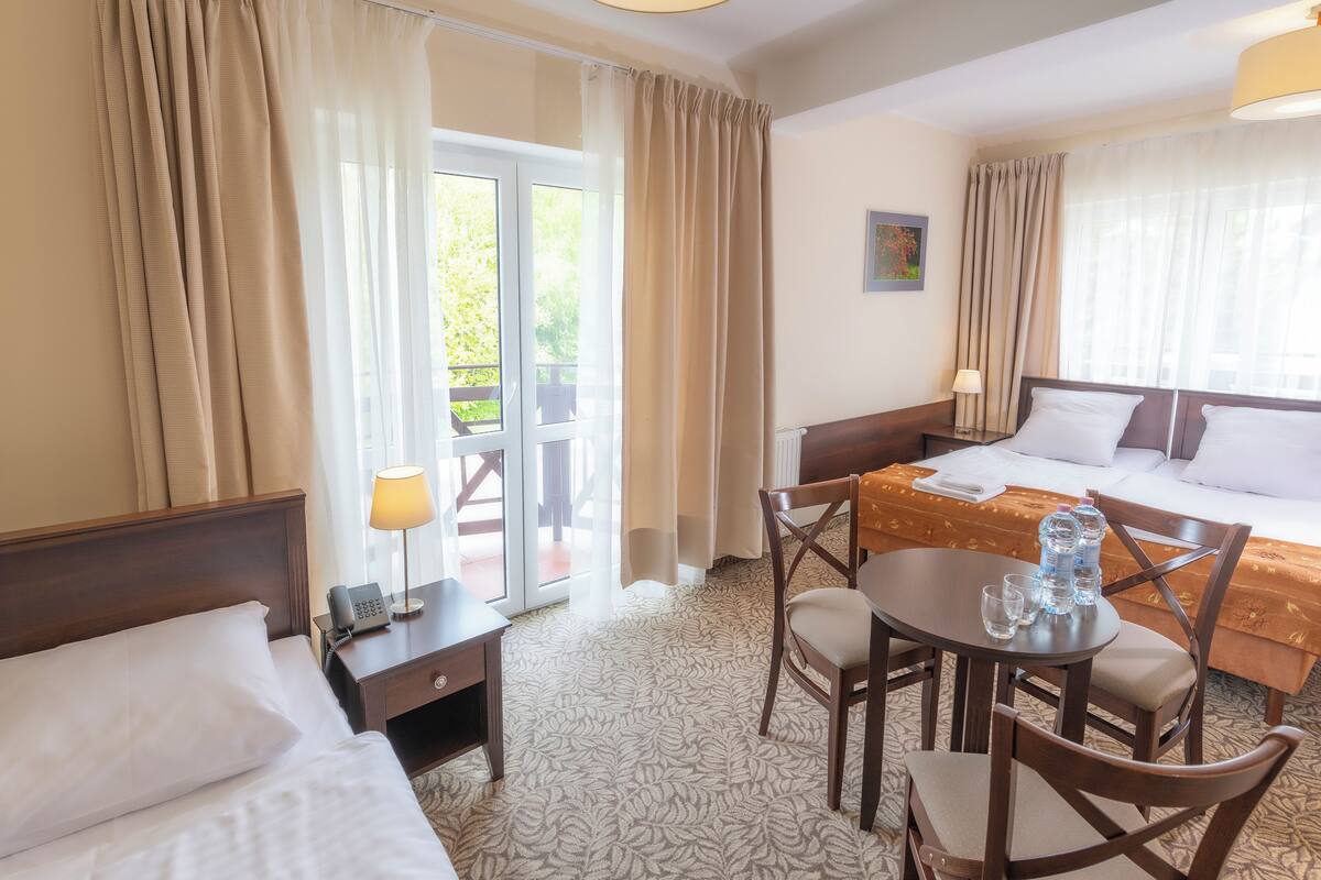 A hotel room with a bed, a desk with a lamp, a table with water bottles, and a view of the outdoors.
