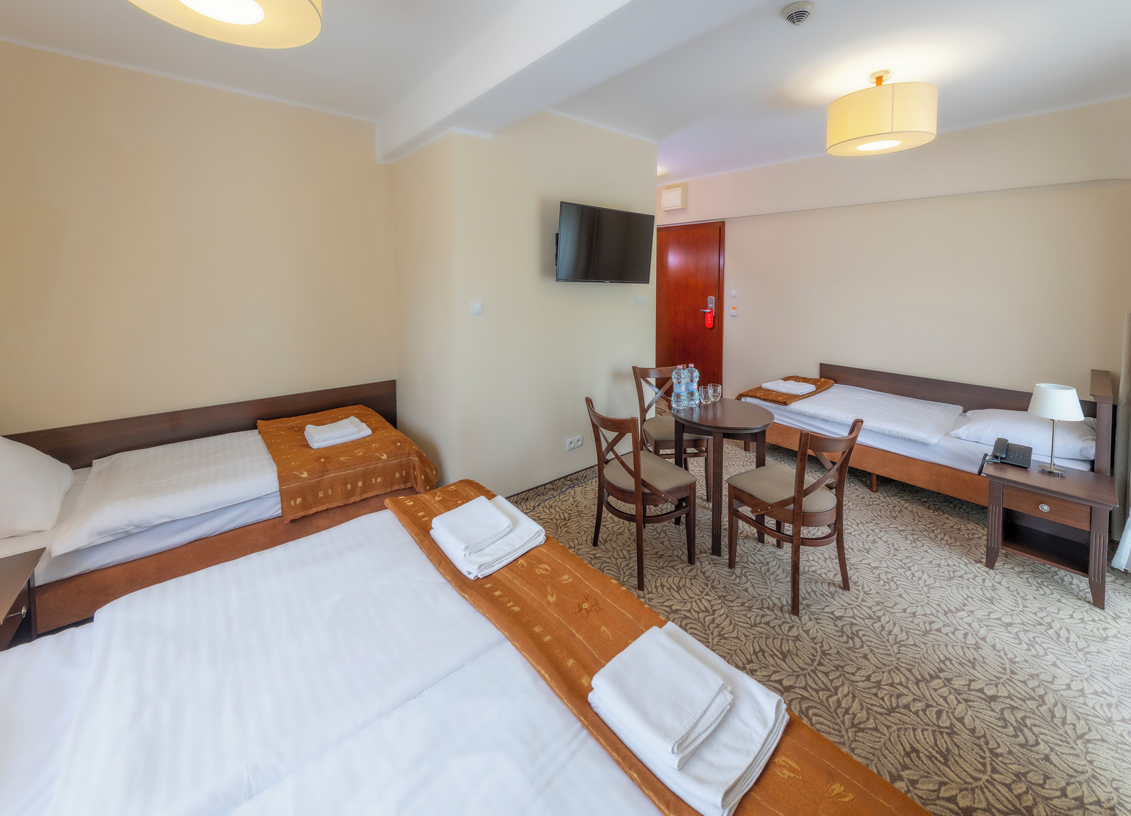 A hotel room with two beds, a small table with chairs, a desk with a lamp, and a TV mounted on the wall.