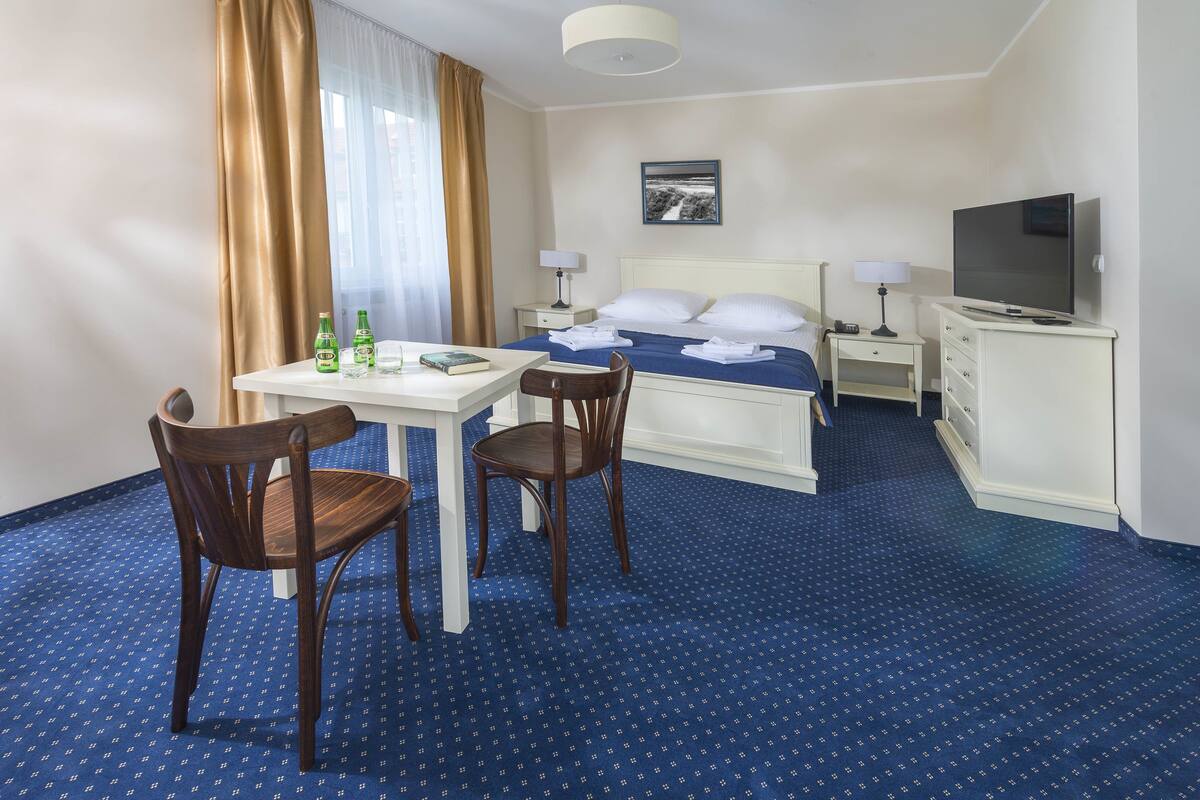 A hotel room with a bed, a TV, a desk with chairs, and a window with curtains.