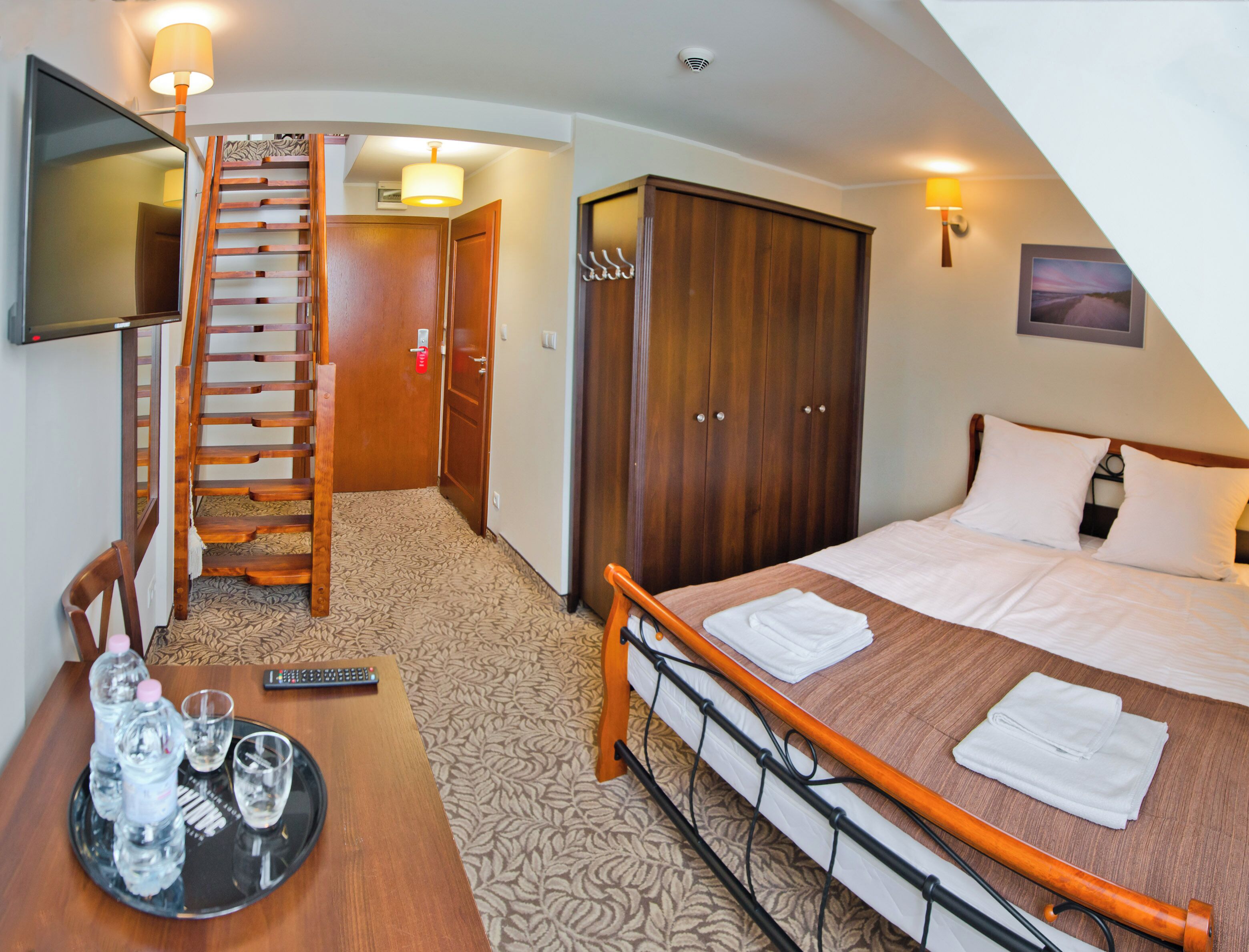 Une chambre d’hôtel comprenant un lit, un bureau avec un plateau de bouteilles d’eau, une télécommande, un téléviseur fixé au mur, une armoire en bois et un escalier.