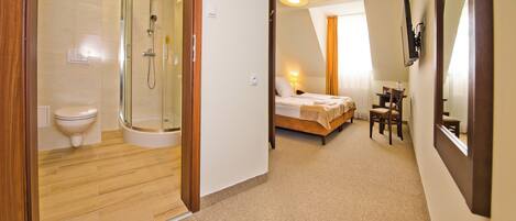 A hotel room with a shower, toilet, bed, and a small table with a chair.