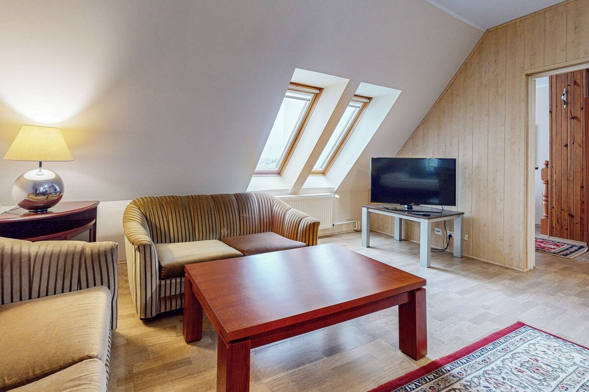 A living room with a sofa, armchair, table, lamp, TV, and a view of the sky through windows.