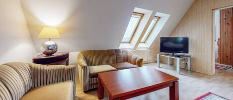 Ein Wohnzimmer mit Sofa, Sessel, Tisch, Lampe, Fernseher und Blick auf den Himmel durch Fenster.