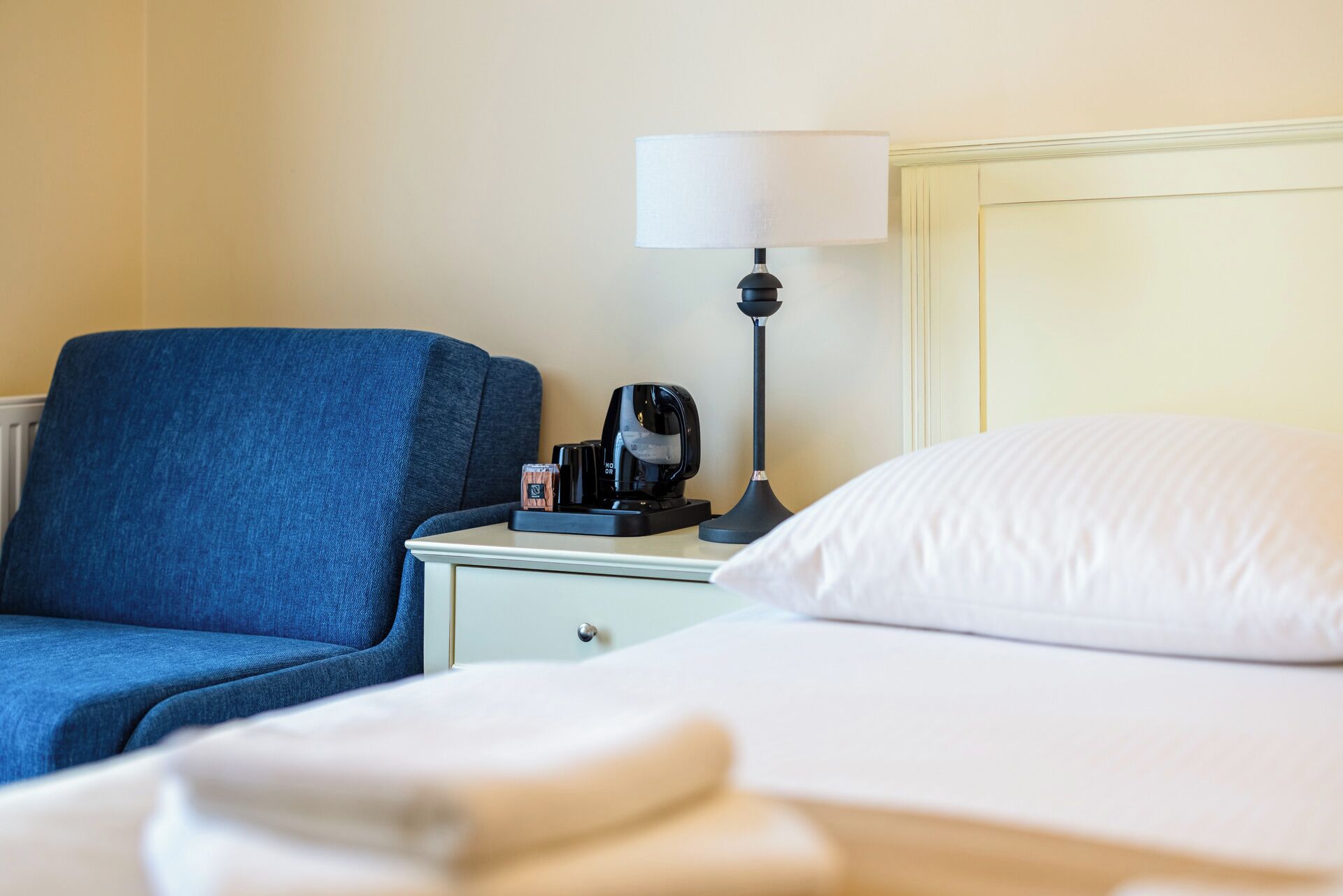A hotel room with a bed, a nightstand, a lamp, a coffee maker, and a blue chair.