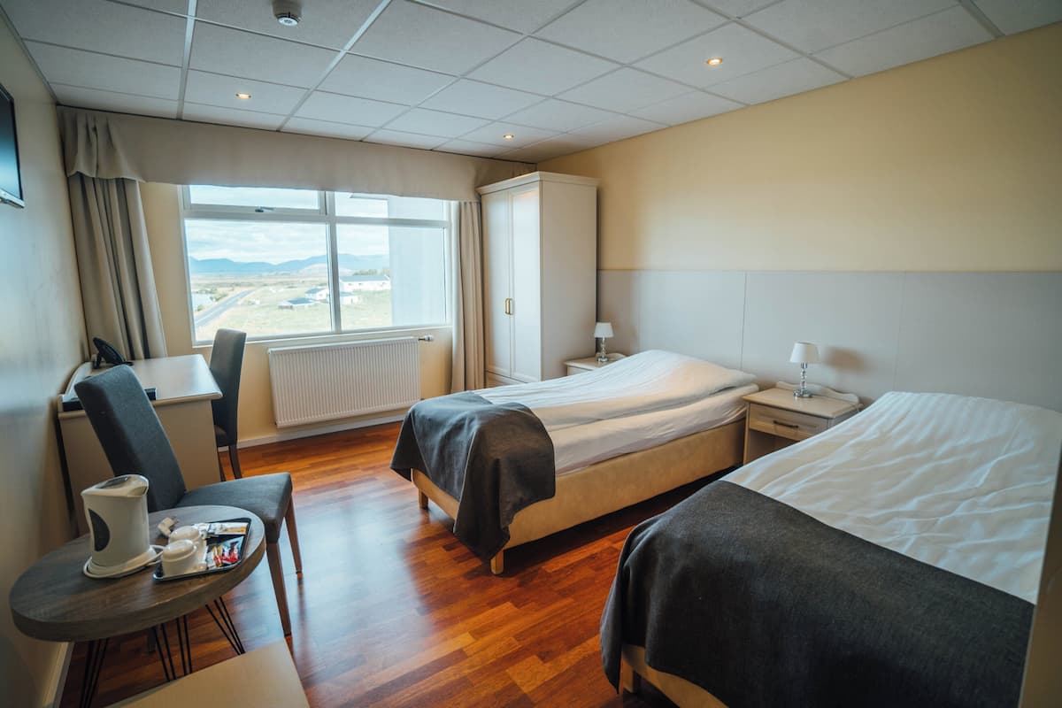 Habitación de hotel con dos camas, un escritorio con silla, un radiador y una ventana con vistas al paisaje.