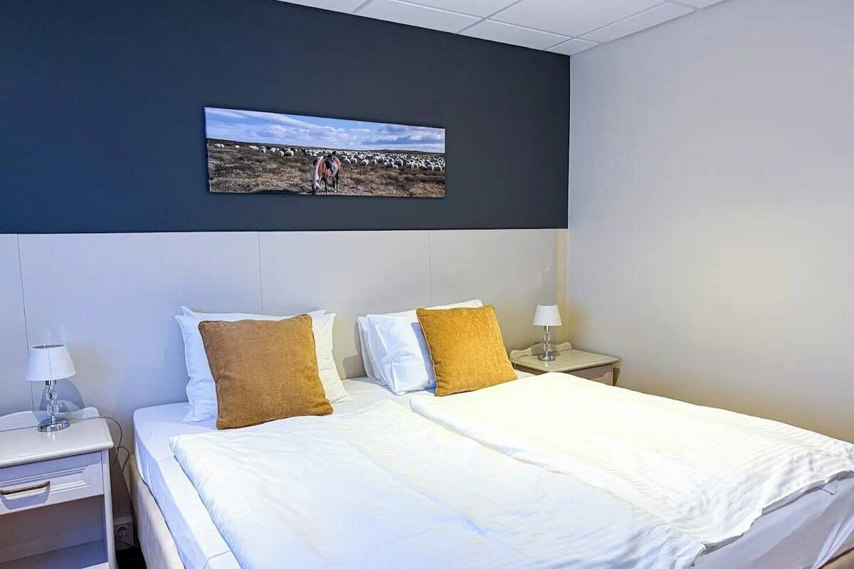 Una habitación de hotel con una cama, dos mesitas de noche, una lámpara y un cuadro de un paisaje.