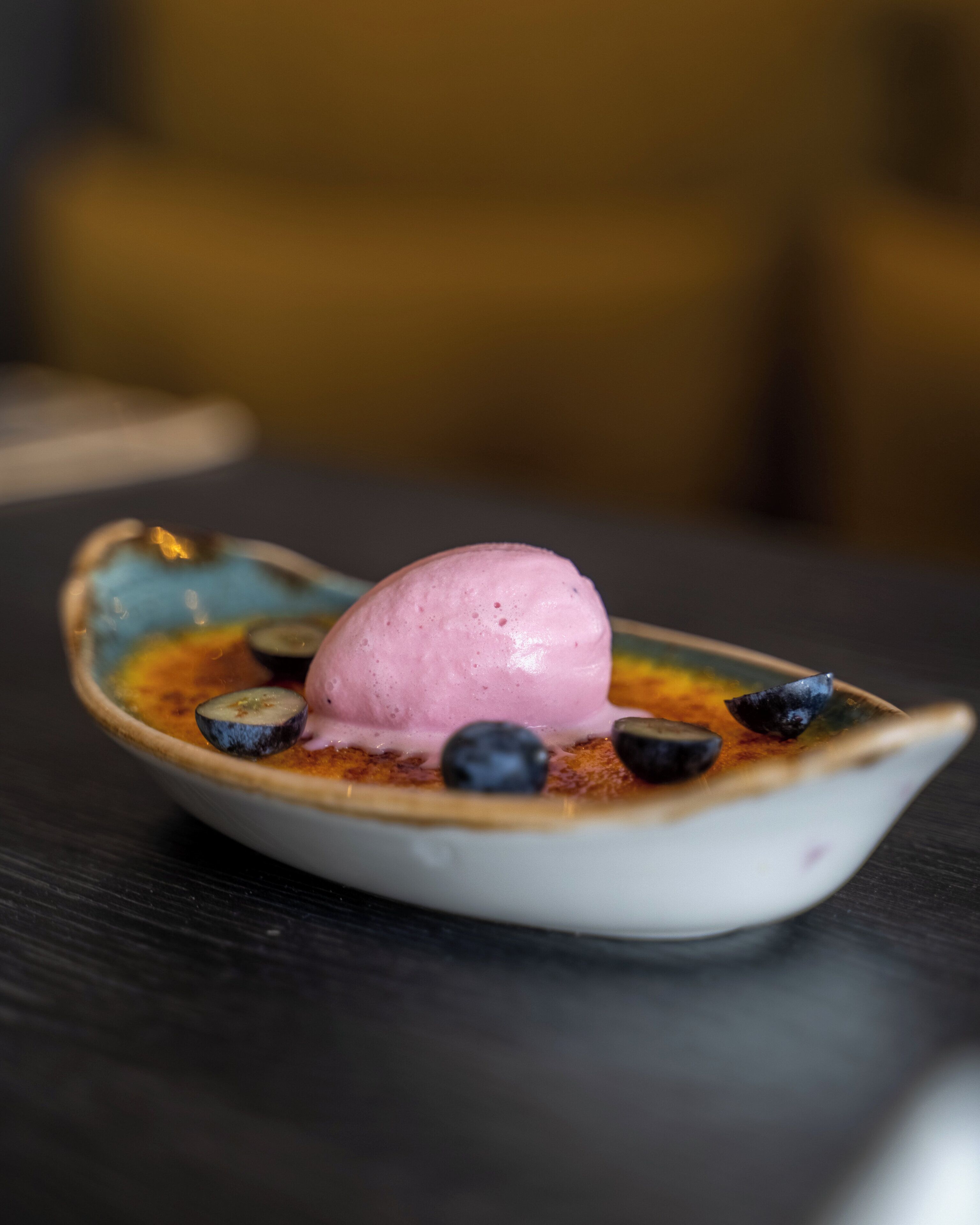 A dessert in a unique boat-shaped dish with a scoop of pink ice cream and blueberries on a dark table.