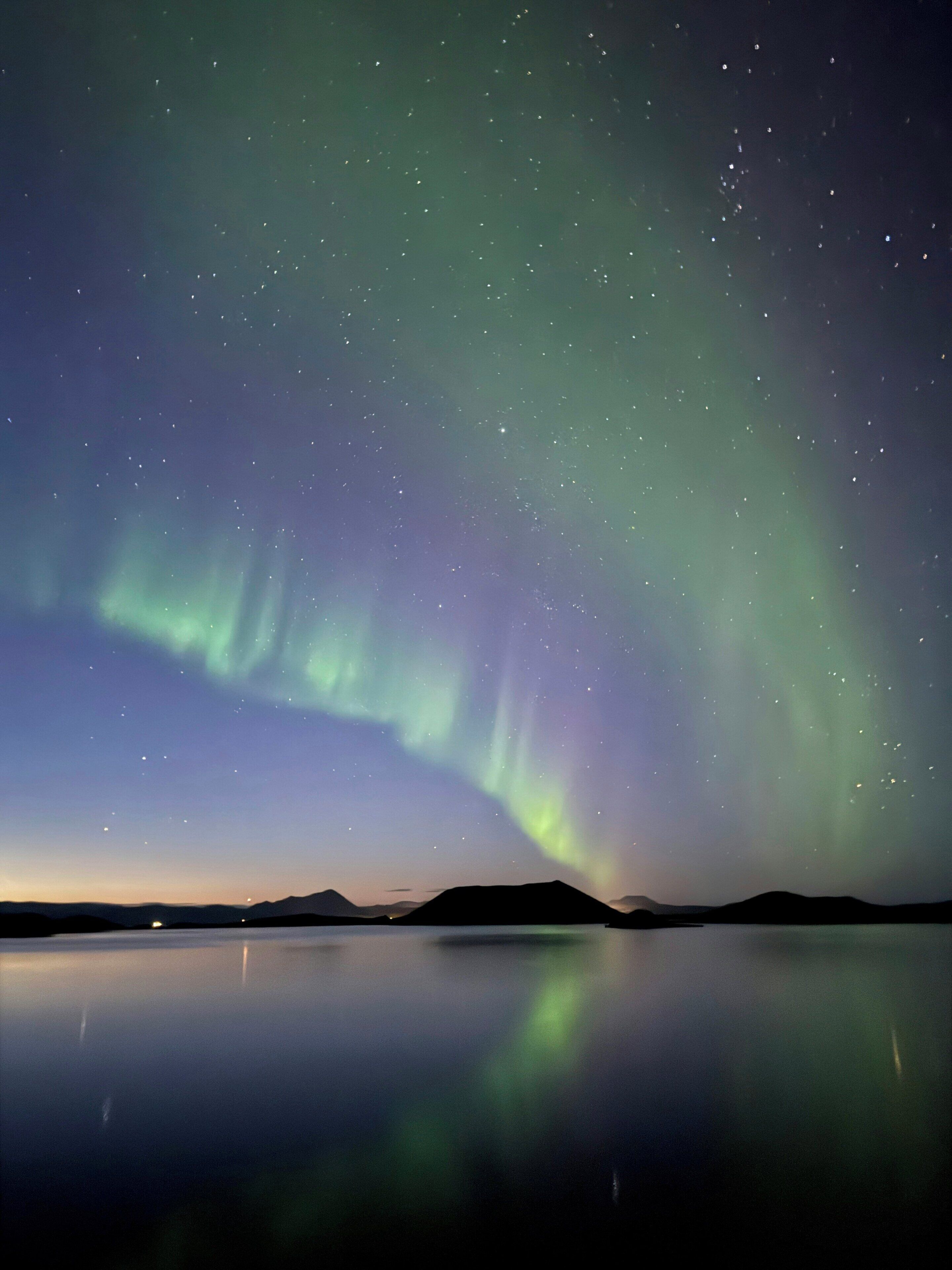 Een nachtelijke hemel met het noorderlicht boven een kalm meer, met bergen op de achtergrond.