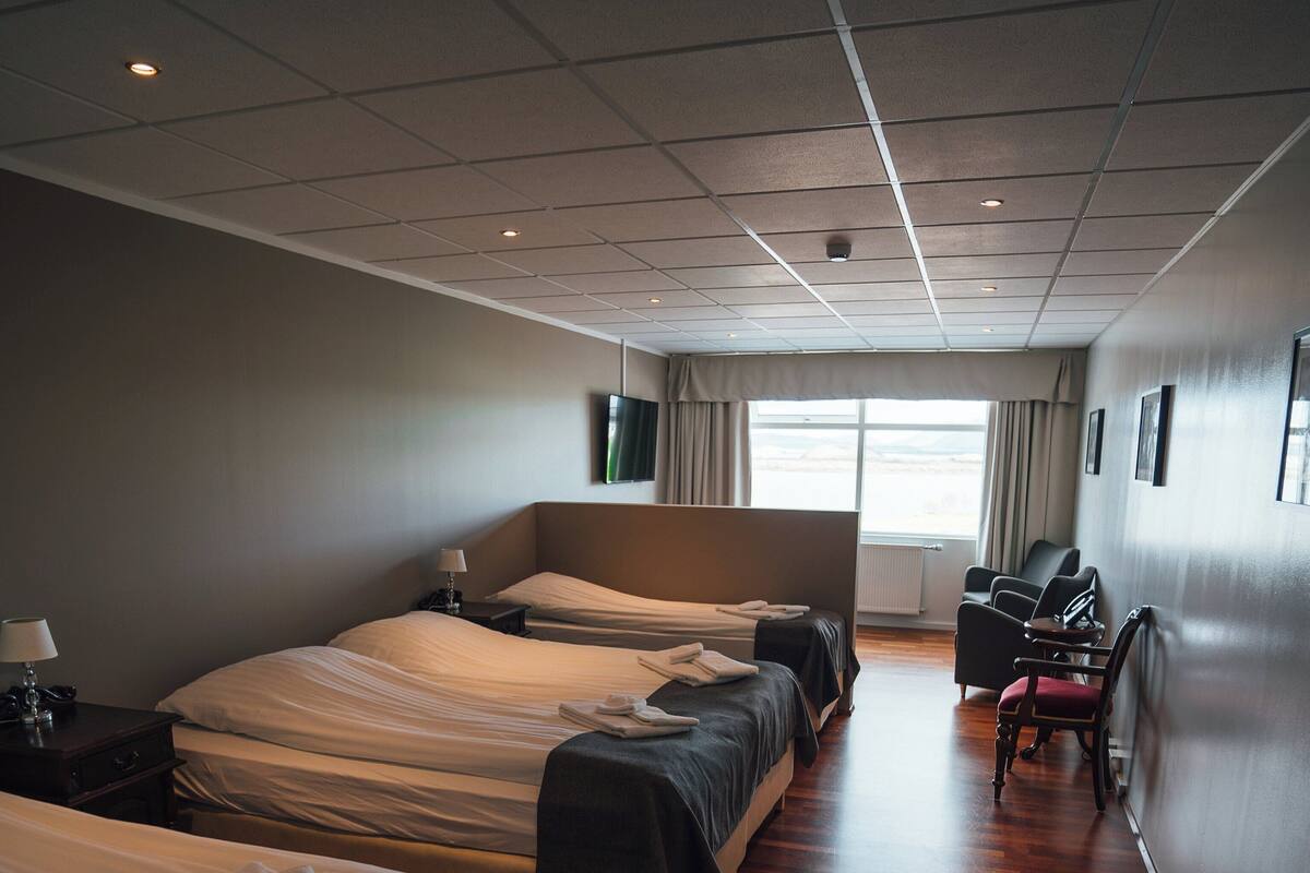 Habitación de hotel con tres camas, un escritorio, una silla y un ventanal con cortinas.