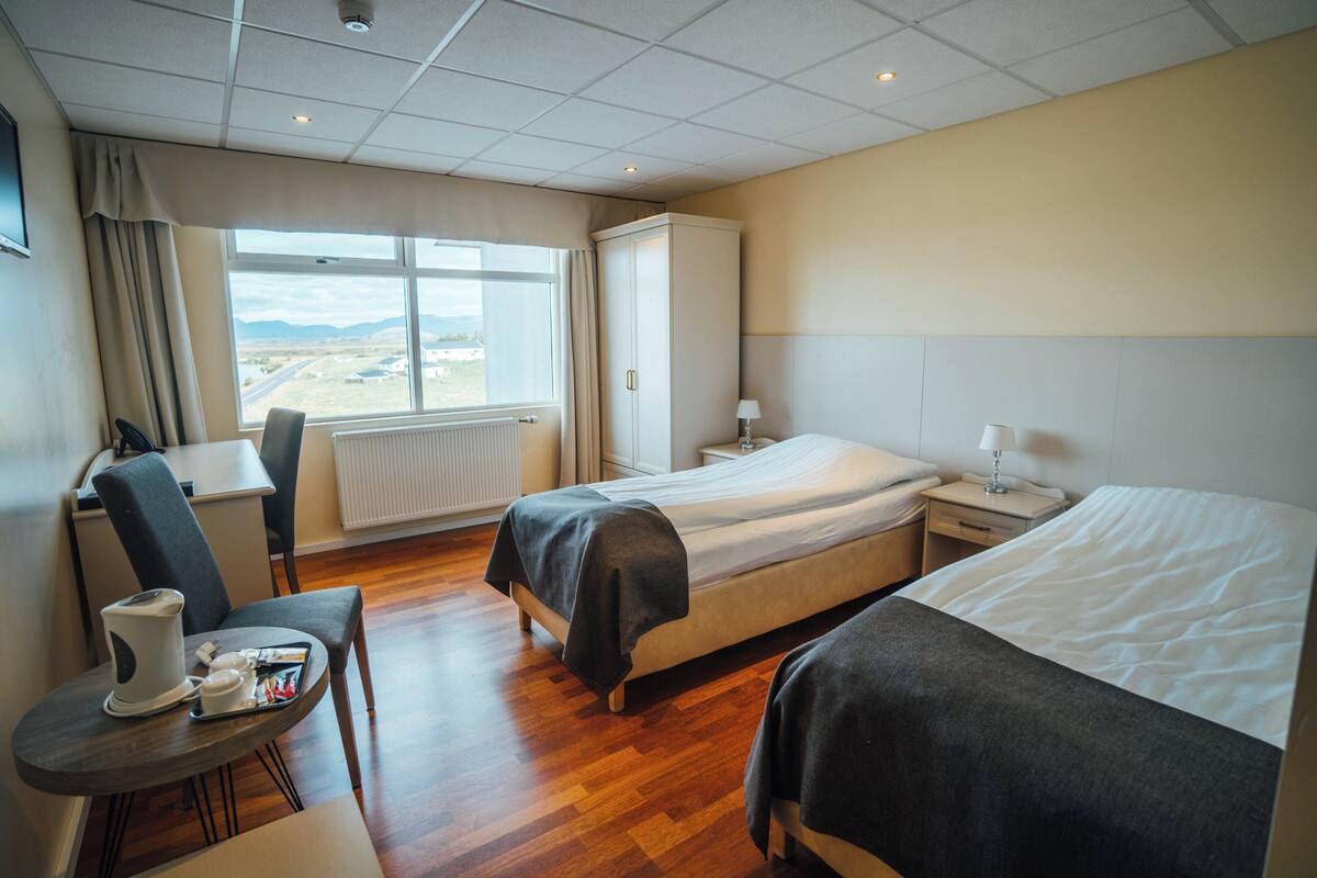 A hotel room with two beds, a desk with a chair, a radiator, and a window with a view of the landscape.