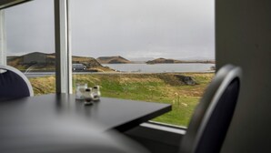 Uma mesa com um copo e uma garrafa, uma cadeira, e vista para uma paisagem com colinas e um corpo d'água pela janela.