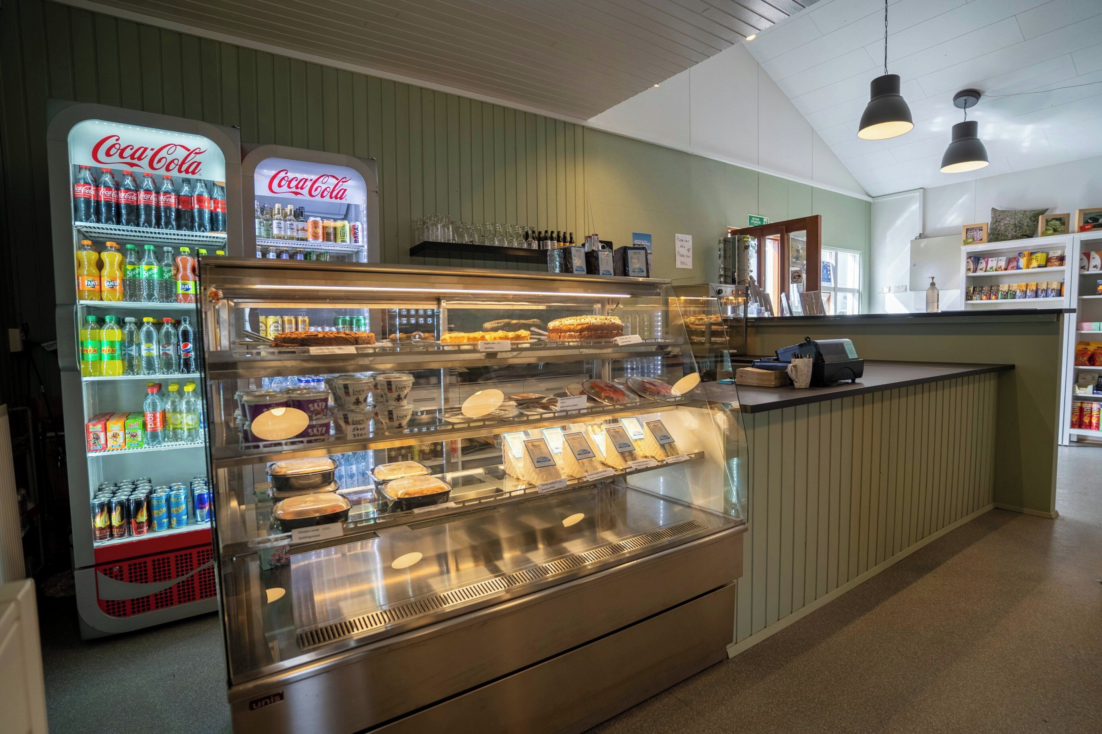 A food display case with various baked goods, a Coca-Cola refrigerator stocked with beverages, and a counter with a cash register.