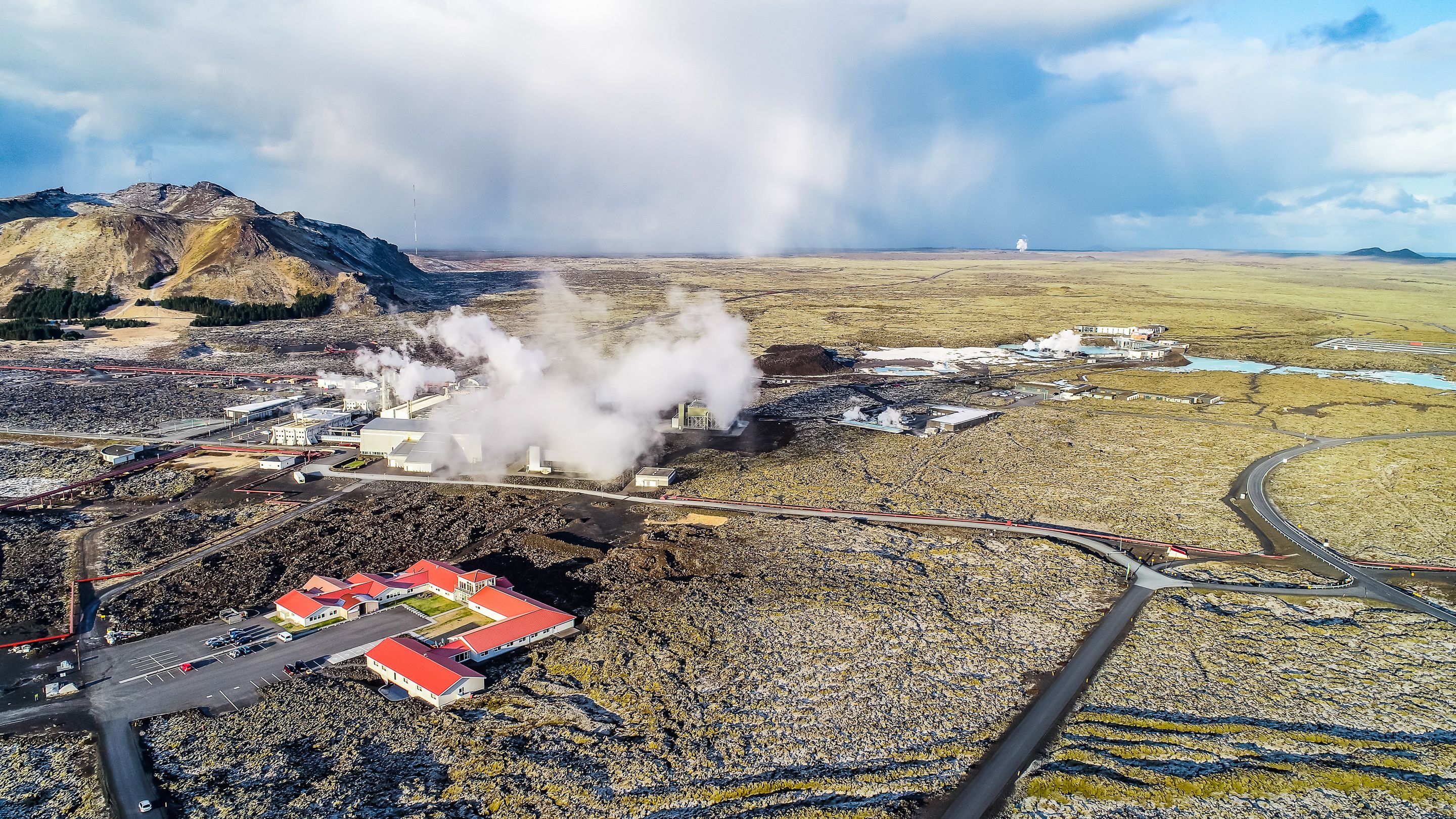An industrial facility with multiple buildings, a parking area, and a mountainous backdrop.