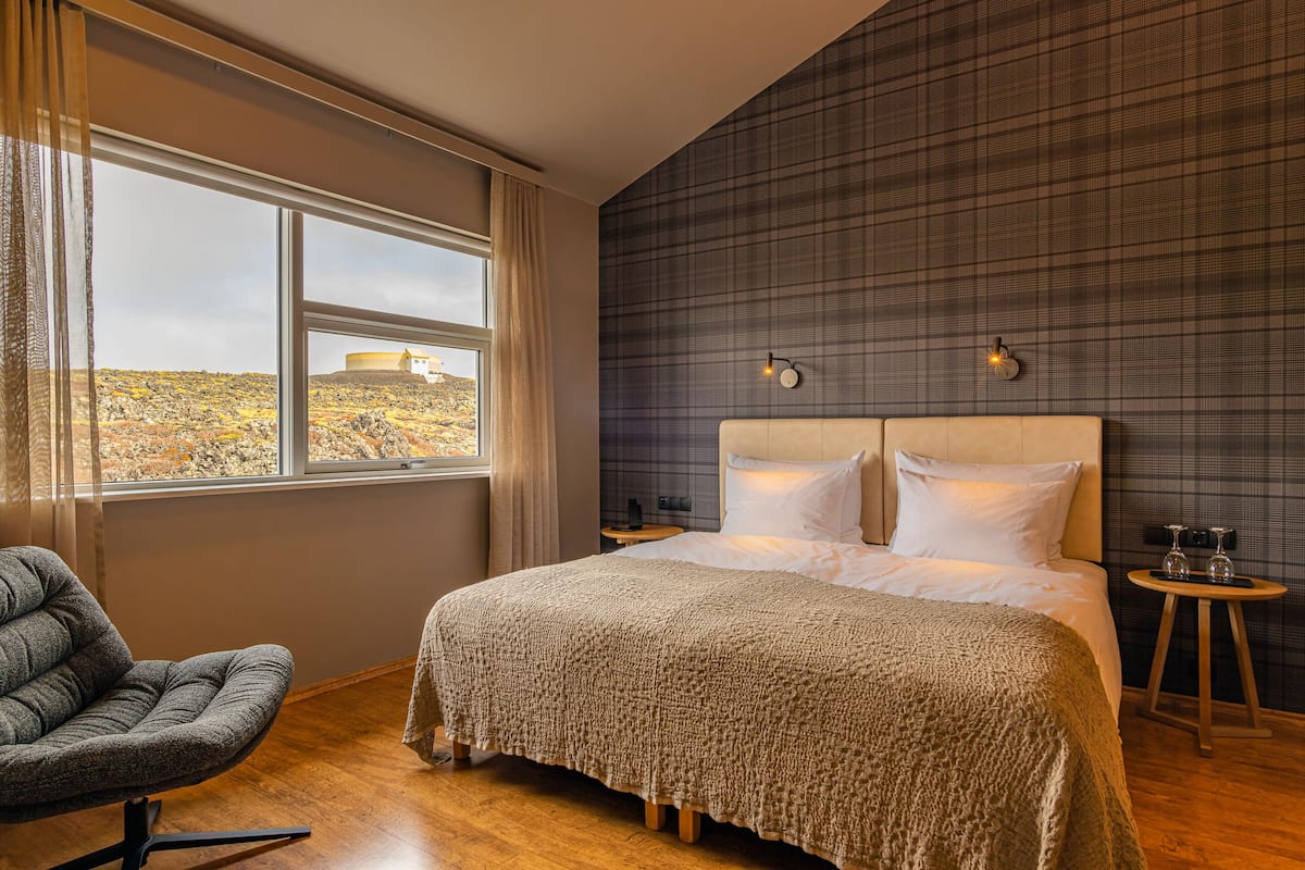 Une chambre avec un grand lit, une chaise, une table de chevet et une vue sur une colline par la fenêtre.