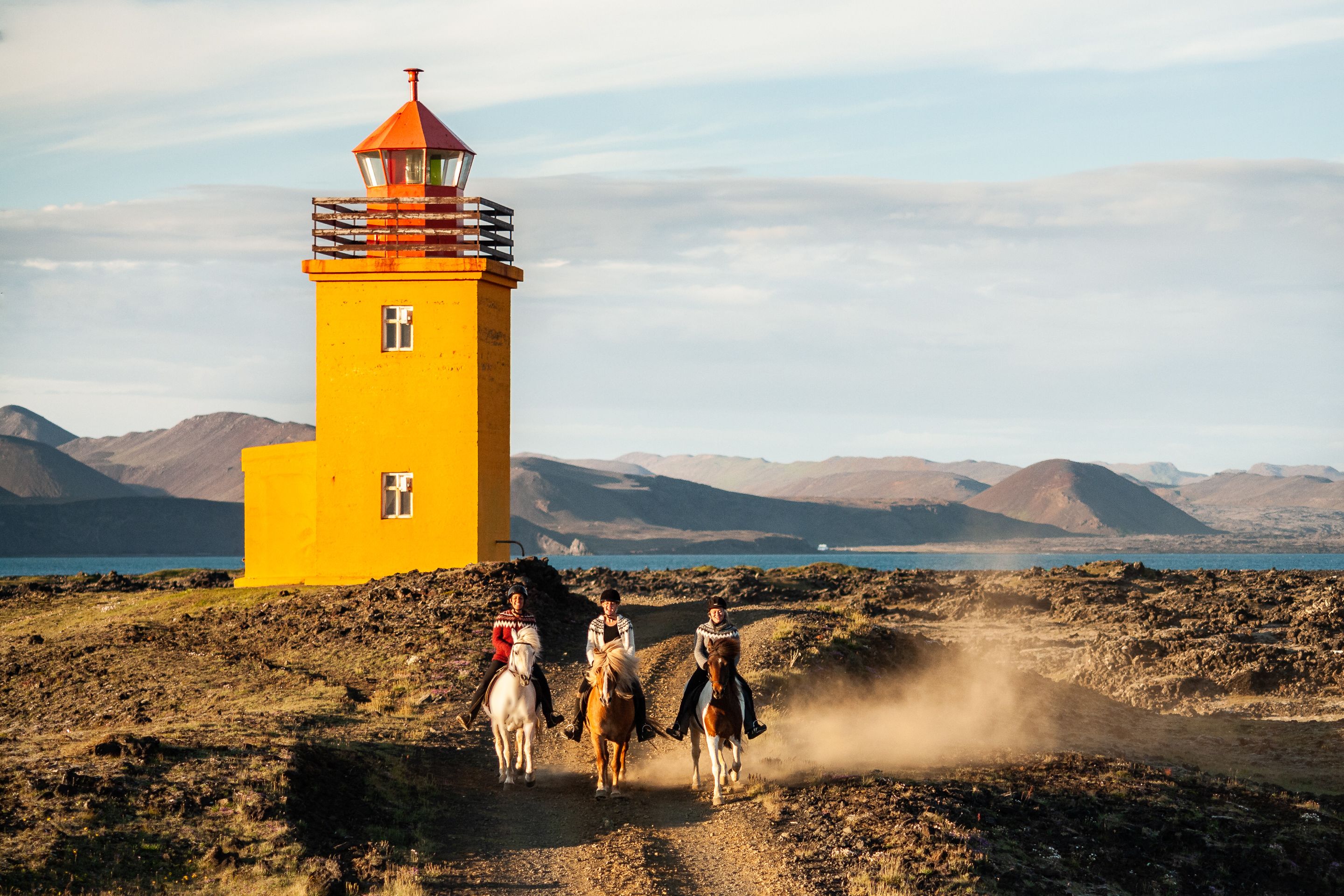 A yellow lighthouse with a red top, surrounded by a rocky landscape and mountains in the background.