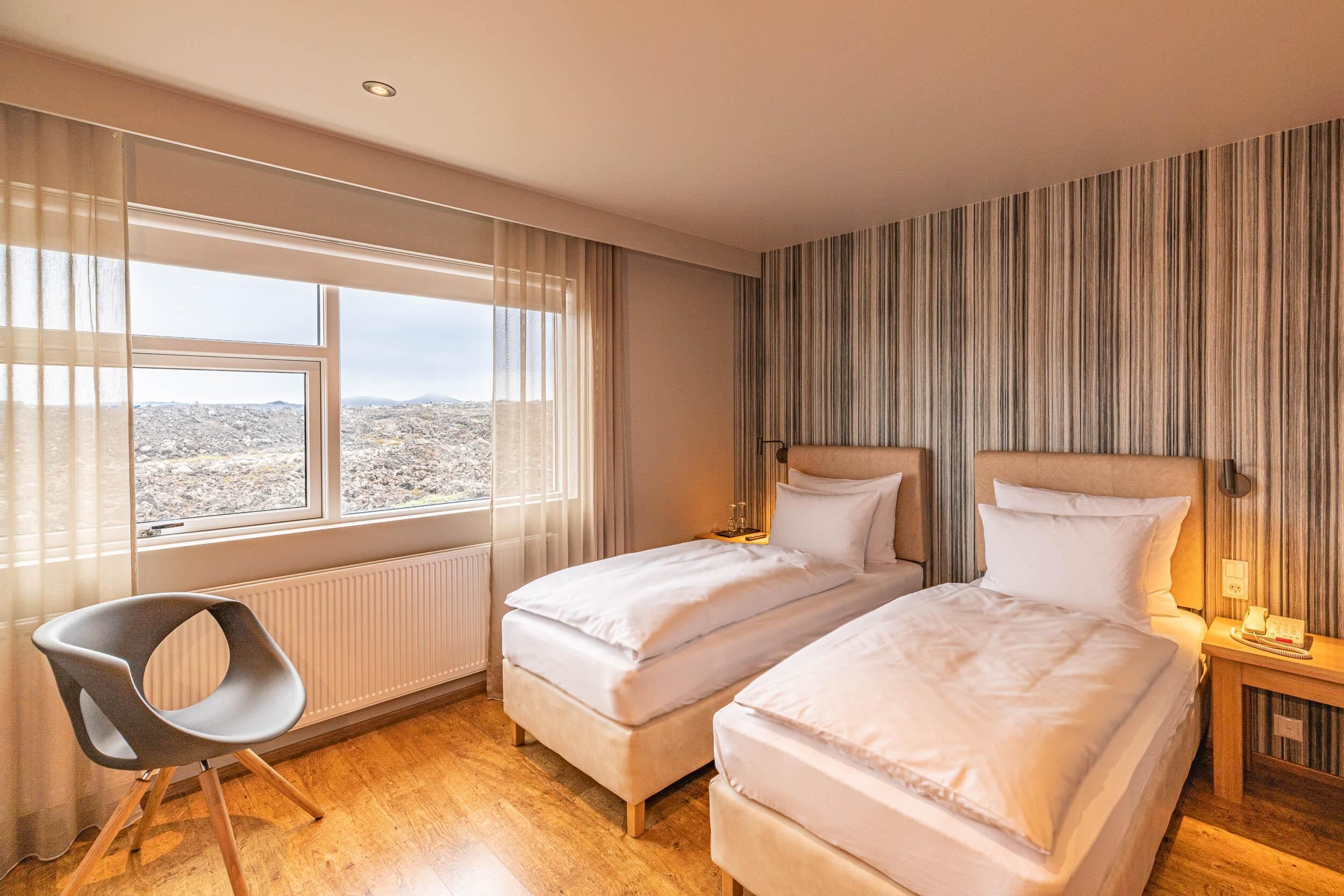 Ein Hotelzimmer mit zwei Betten, einem Sessel, einem Fenster mit Aussicht und einer gestreiften Wand.