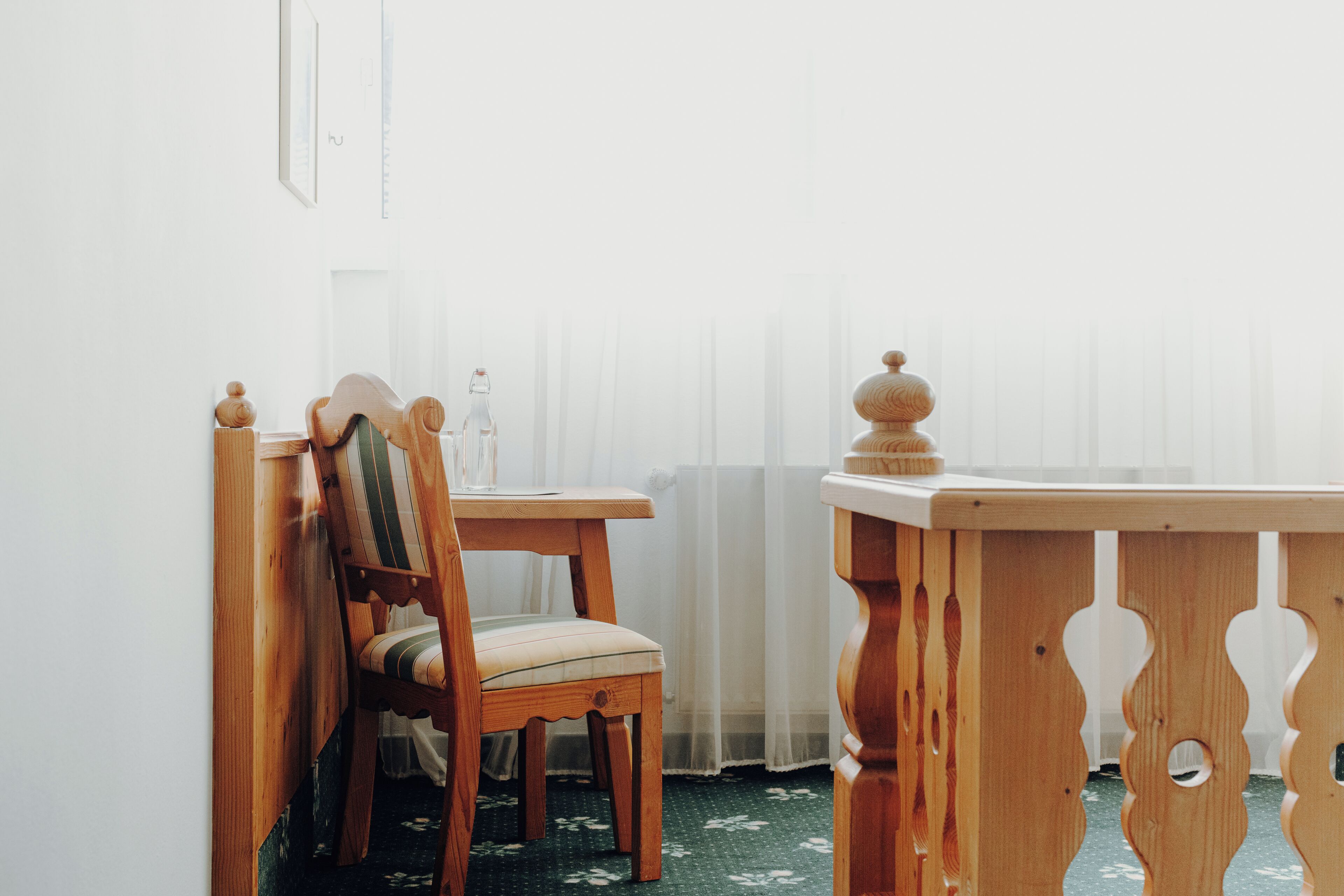Une chaise et une table en bois se trouvent dans une pièce dont un mur est blanc et qui possède une fenêtre ornée de rideaux légers.