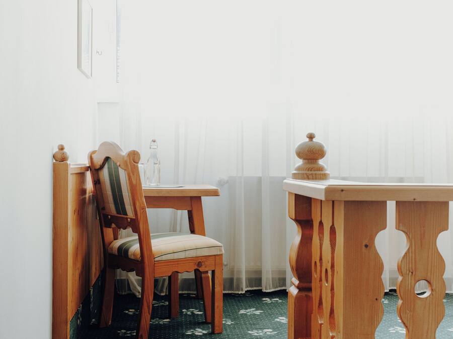 Une chaise et une table en bois se trouvent dans une pièce dont un mur est blanc et qui possède une fenêtre ornée de rideaux légers.