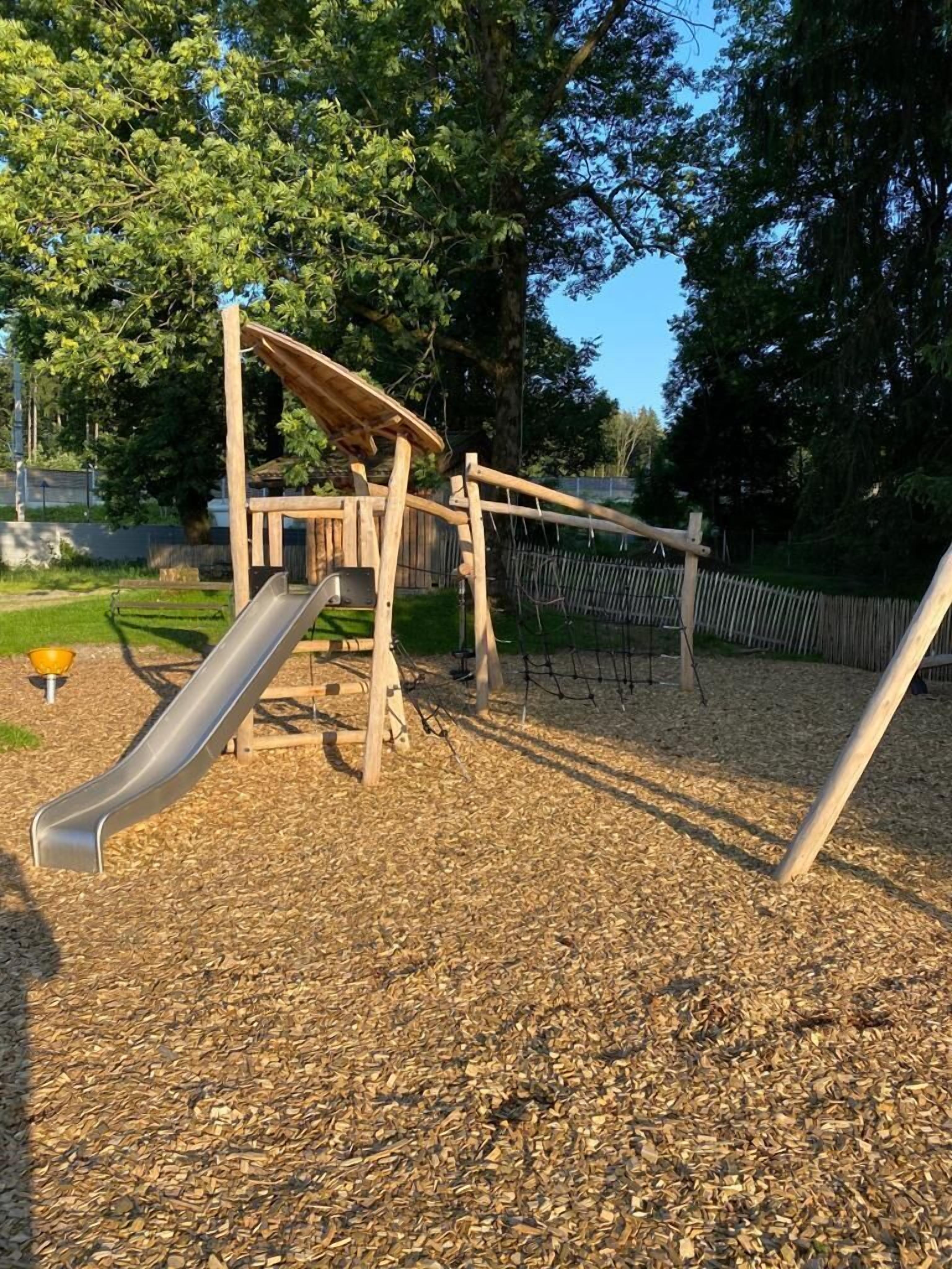 Un parque infantil con tobogán y columpios, rodeado de árboles y una cerca.