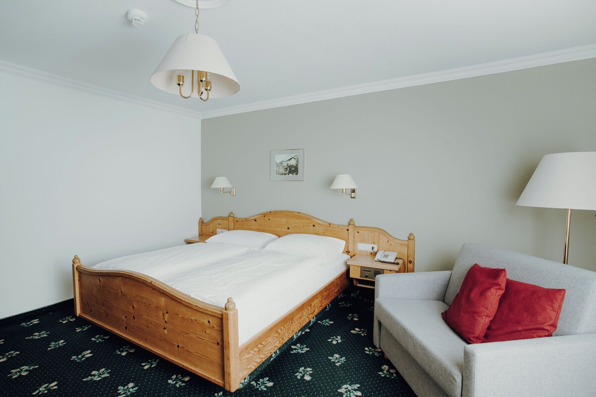 A neatly arranged hotel room with a wooden bed, a grey sofa, and a red pillow.