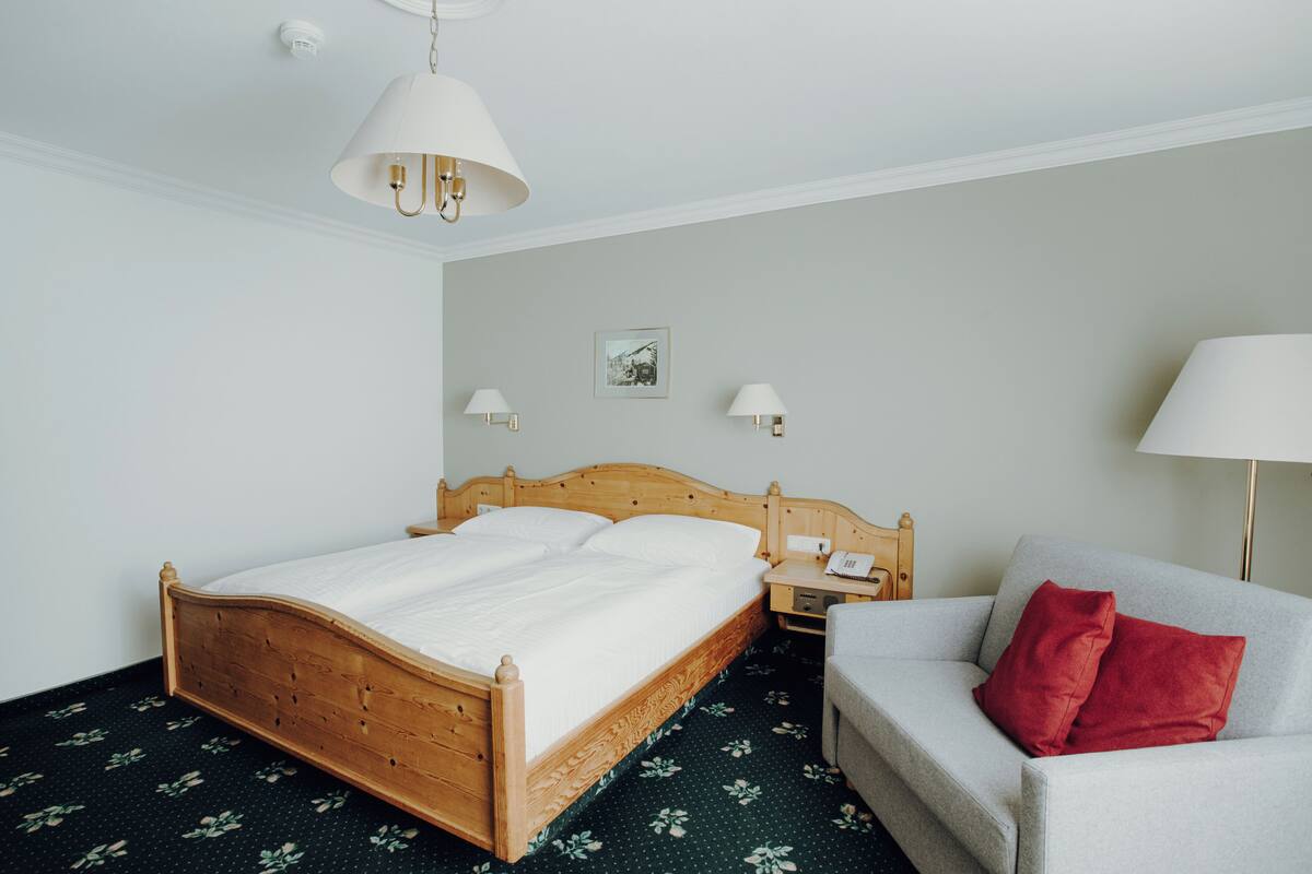 A neatly arranged hotel room with a wooden bed, a grey sofa, and a red pillow.