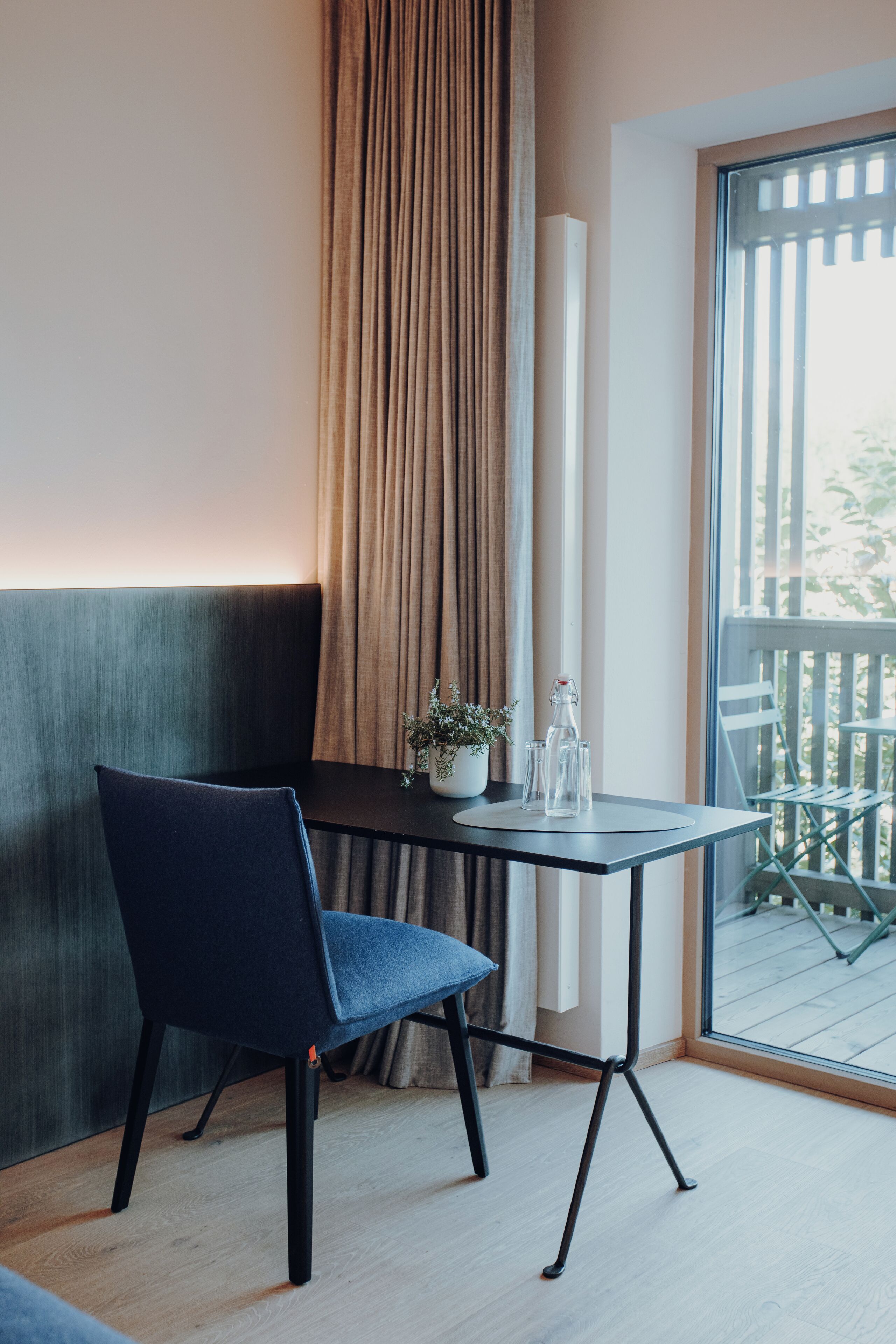 A modern room with a dark blue chair, a small table with a potted plant, and a view of a balcony through a large window.