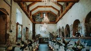 An interior with a high ceiling and exposed wooden beams, a central aisle with white chairs, and chandeliers. - Belleek Castle (Ballina)