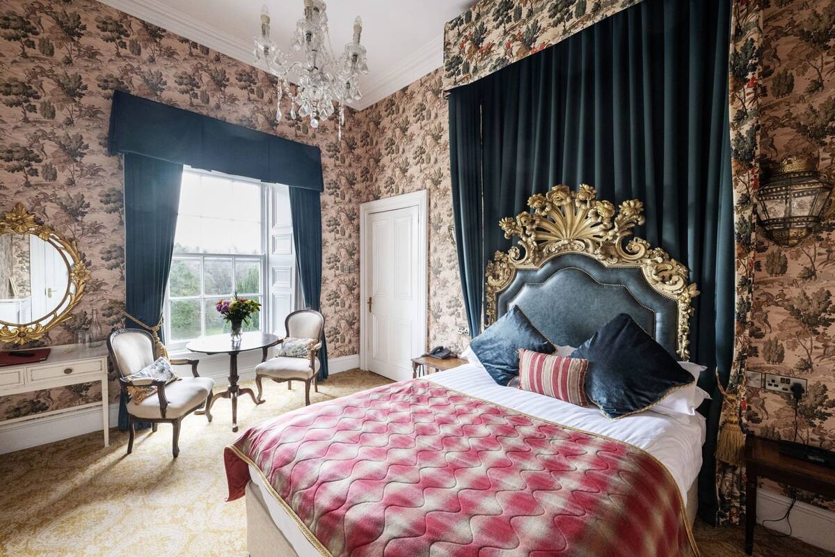 A bedroom with a large bed, a chandelier, a desk with a chair, a window, and floral wallpaper.