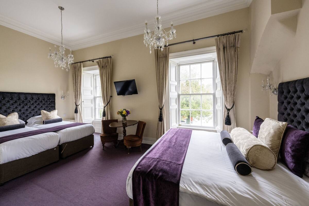 A hotel room with two beds, a TV, a chandelier, and a window with curtains.