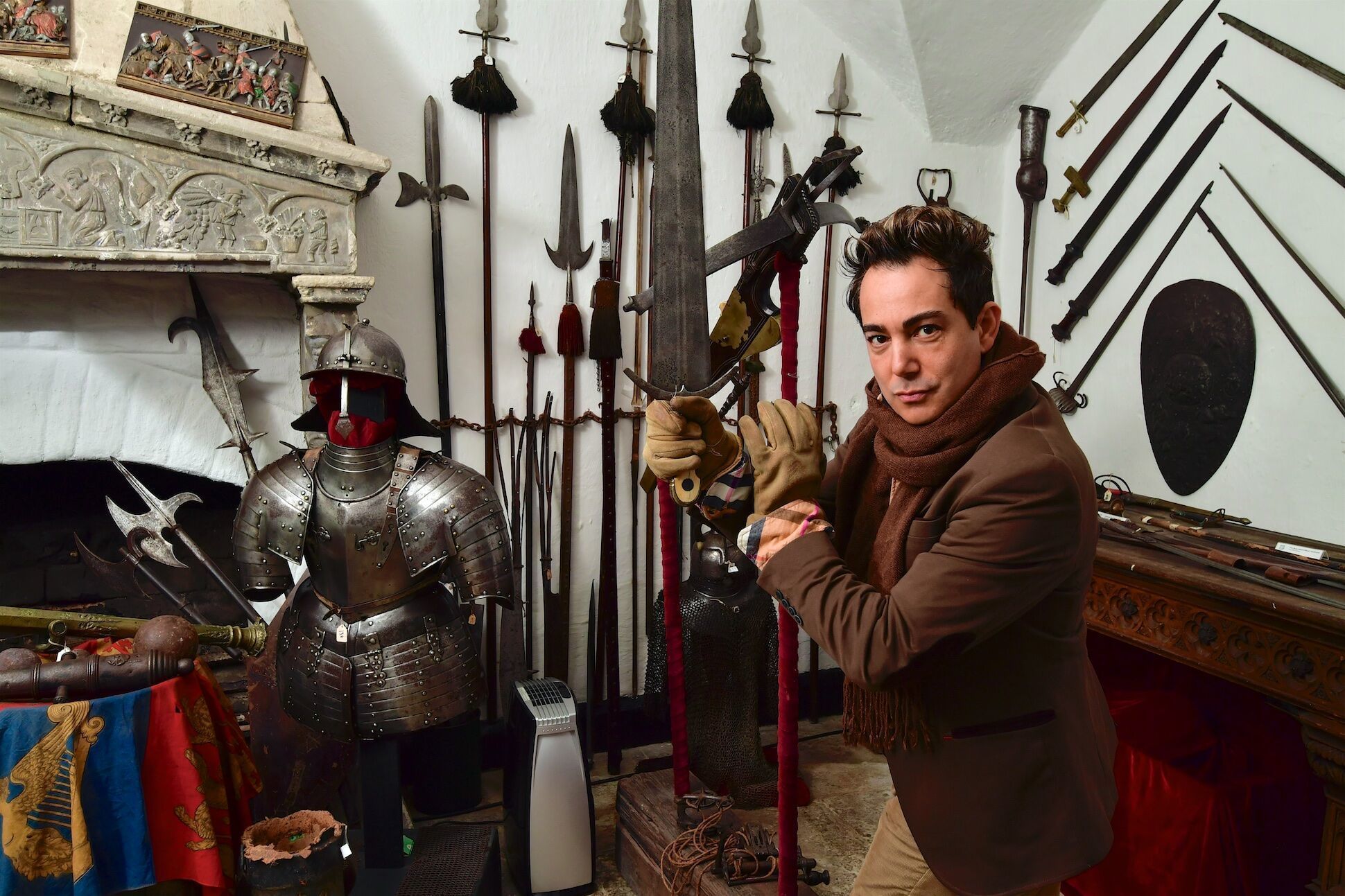 A room with a medieval knight suit of armor, various weapons, and a tapestry.