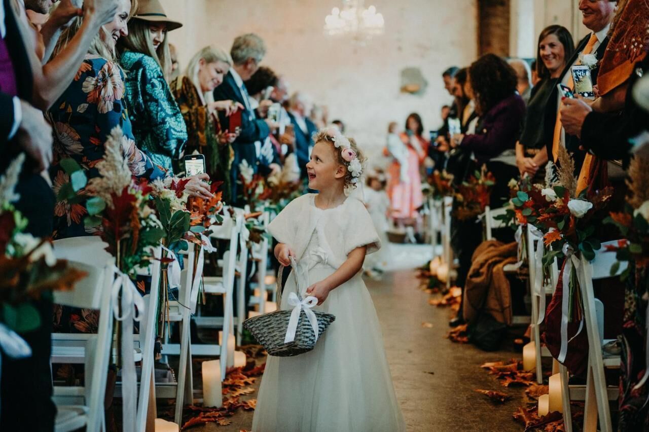 Eine Hochzeitszeremonie mit Gästen, die auf weißen Stühlen sitzen, einer Braut im weißen Kleid, die einen Korb hält, und floralen Dekorationen.