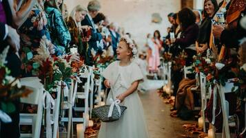 Eine Hochzeitszeremonie mit Gästen, die auf weißen Stühlen sitzen, einer Braut im weißen Kleid, die einen Korb hält, und floralen Dekorationen.