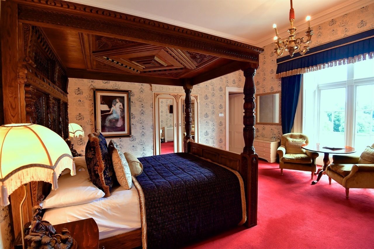 A four-poster bed with a dark quilt, a chandelier, and a red carpeted floor.