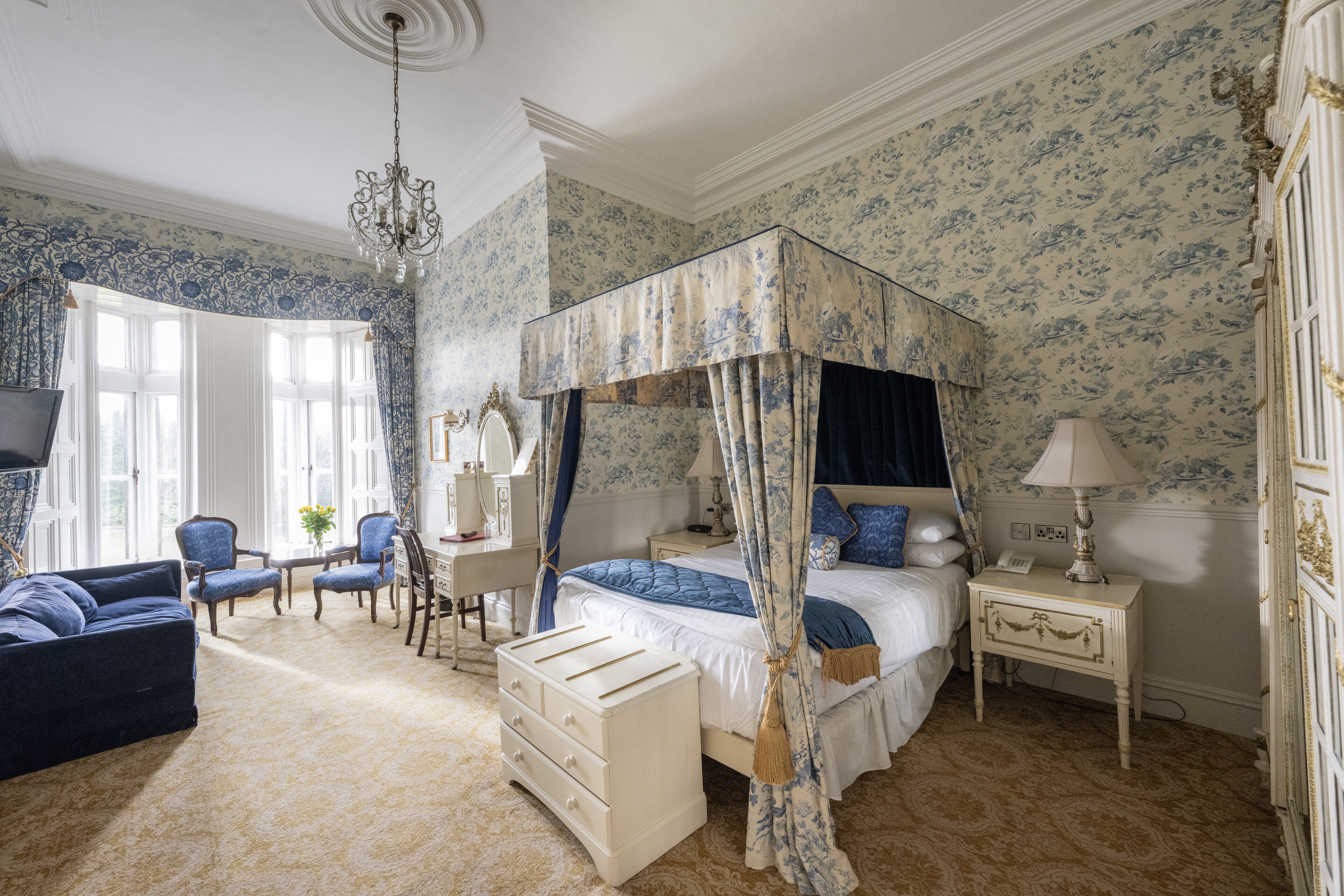 A bedroom with a four-poster bed, a desk with a chair, a sofa, and a nightstand with a lamp.