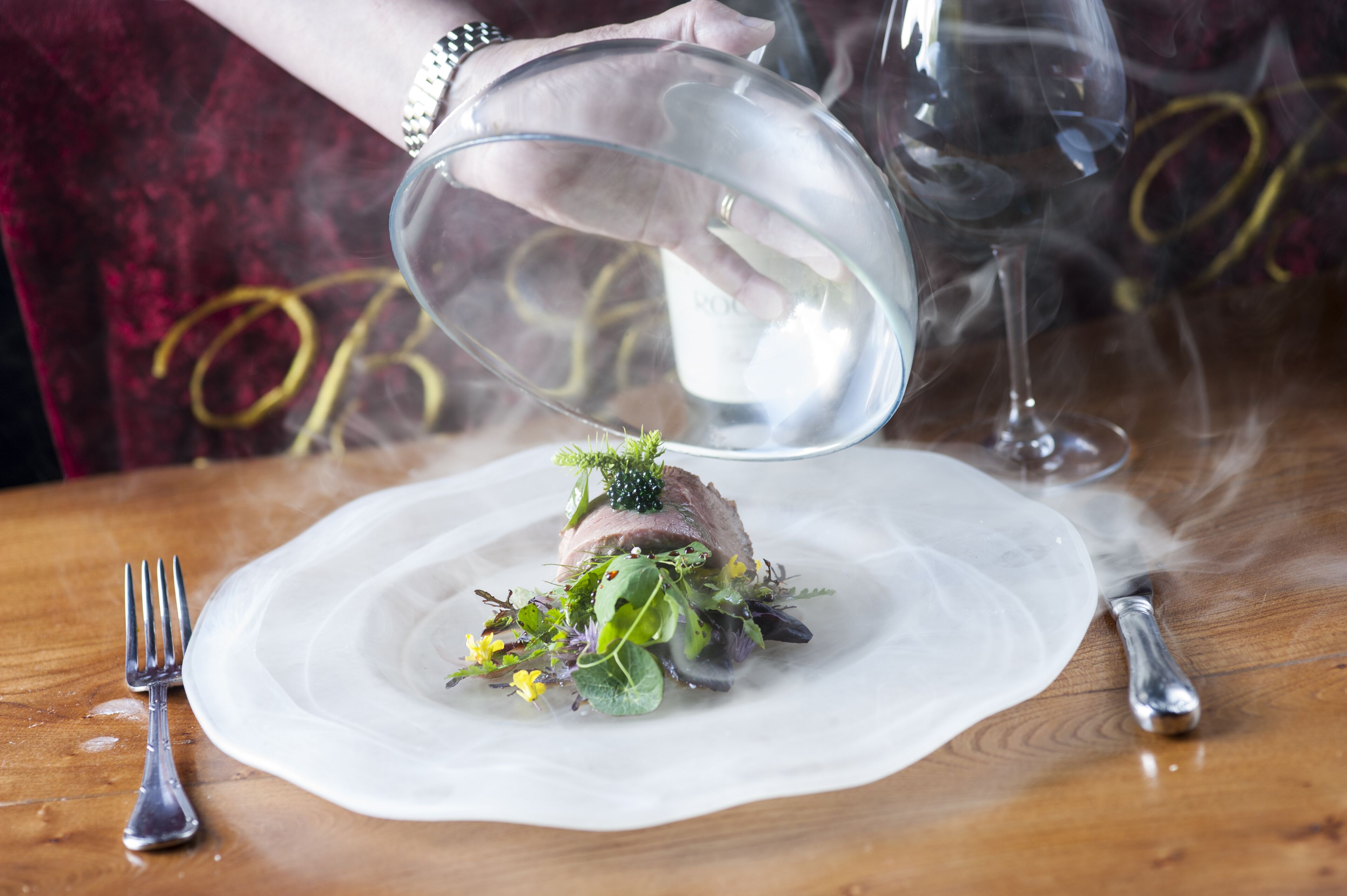 A person pouring a clear liquid over a dish with a meat centerpiece and greens, on a wooden table with a fork and knife.
