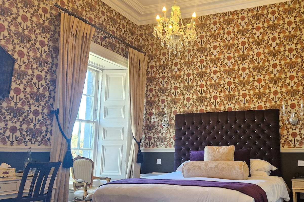 A bedroom with a tufted headboard, floral wallpaper, a chandelier, and a bed with multiple pillows.