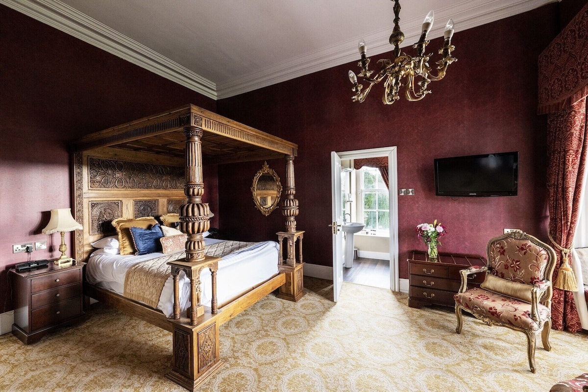 A four-poster bed with a wooden frame, a chandelier, a television, and a floral-patterned armchair.