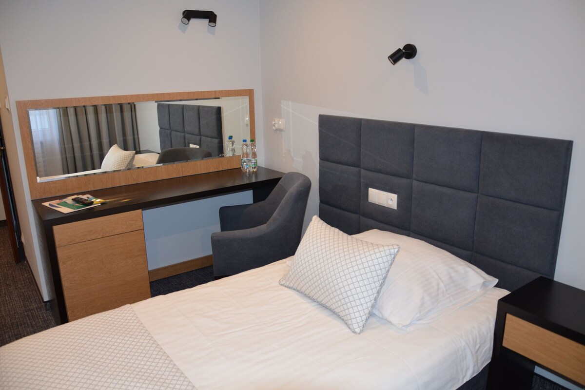 A hotel room with a bed, a desk, a chair, a mirror, and a nightstand.