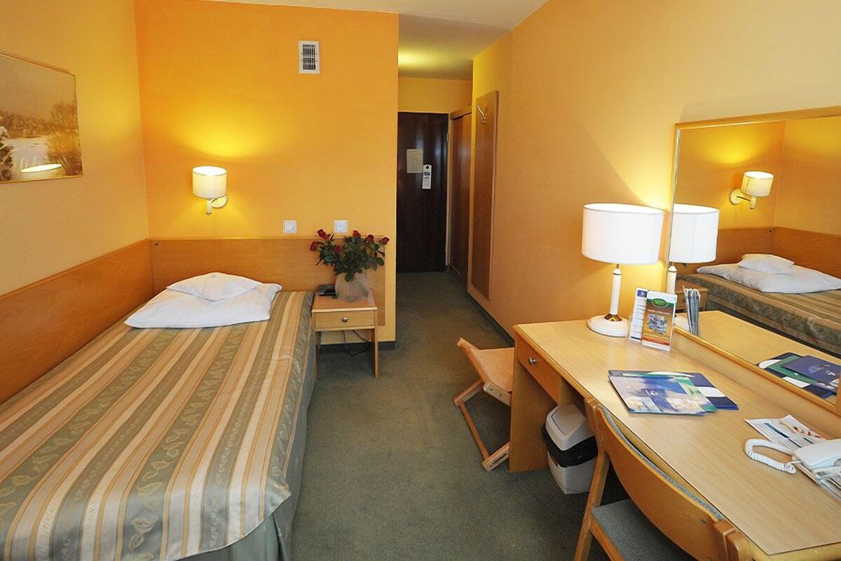A hotel room with two single beds, a desk with a chair, a small table with a vase of flowers, and a large mirror.