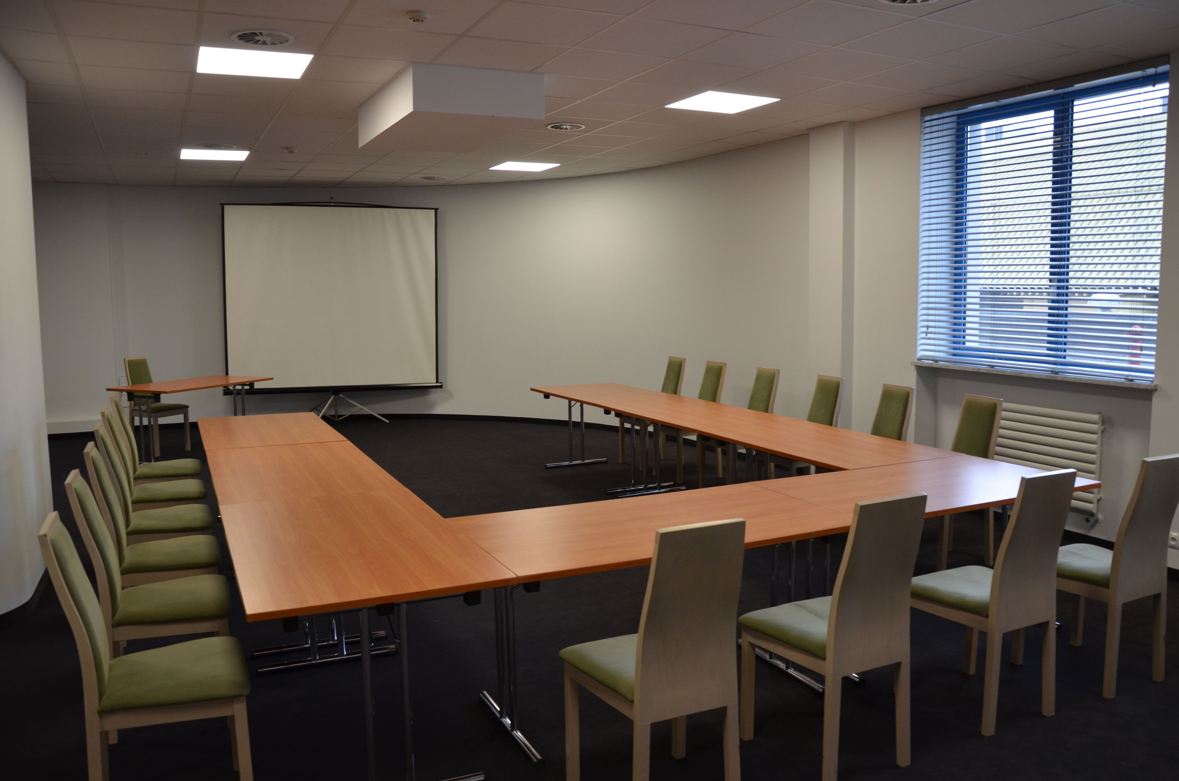 Ett konferensrum med ett långt bord, stolar, en projektorduk och ett fönster med persienner.