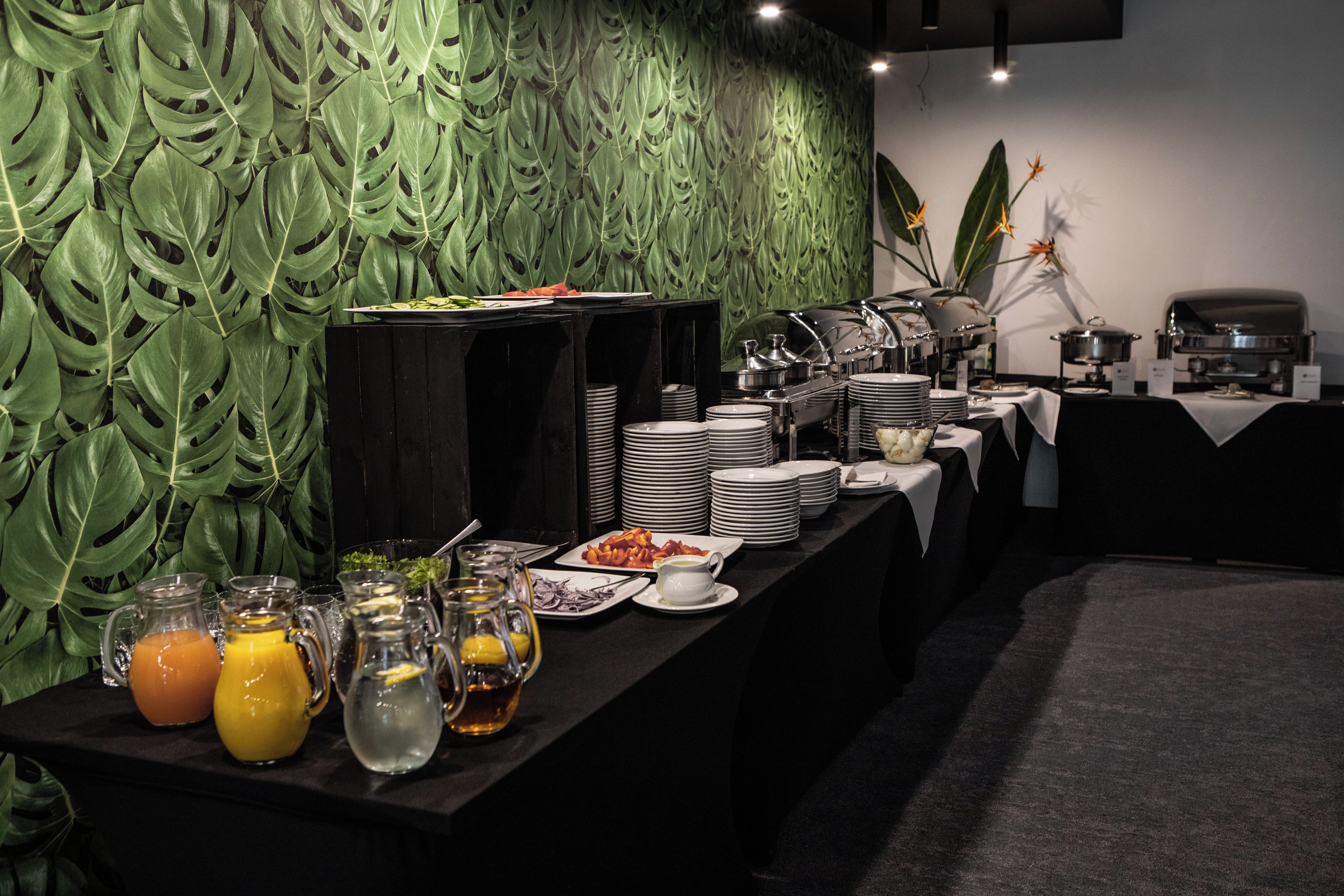 A buffet setup with various food items and beverages, a coffee station, and a decorative green leaf wall.