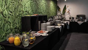 A buffet setup with various food items and beverages, a coffee station, and a decorative green leaf wall.
