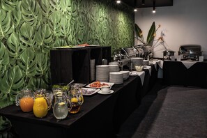 Een buffetopstelling met diverse gerechten en drankjes, een koffiehoek, en een decoratieve wand van groene bladeren.