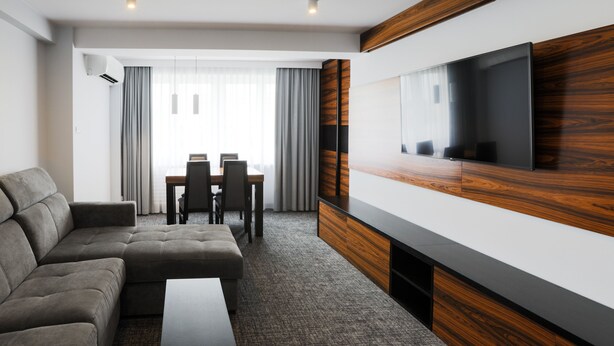 A modern hotel room with a sectional sofa, a flat-screen TV mounted on a wooden panel, a dining area with a table and chairs, and a large window with curtains.