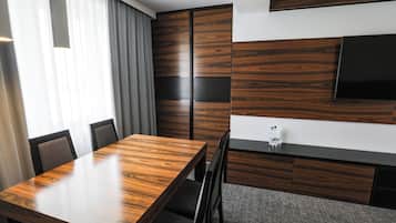 Ett modernt hotellrum med ett matbord i trä, grå stoppade stolar, en platt-TV som är monterad på väggen och ett stort fönster med tunna, genomskinliga gardiner.