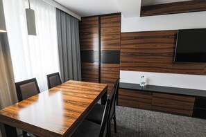 Habitación de hotel moderna con una mesa de comedor de madera, sillas tapizadas de color gris, un televisor de pantalla plana montado en la pared y un ventanal con cortinas translúcidas.