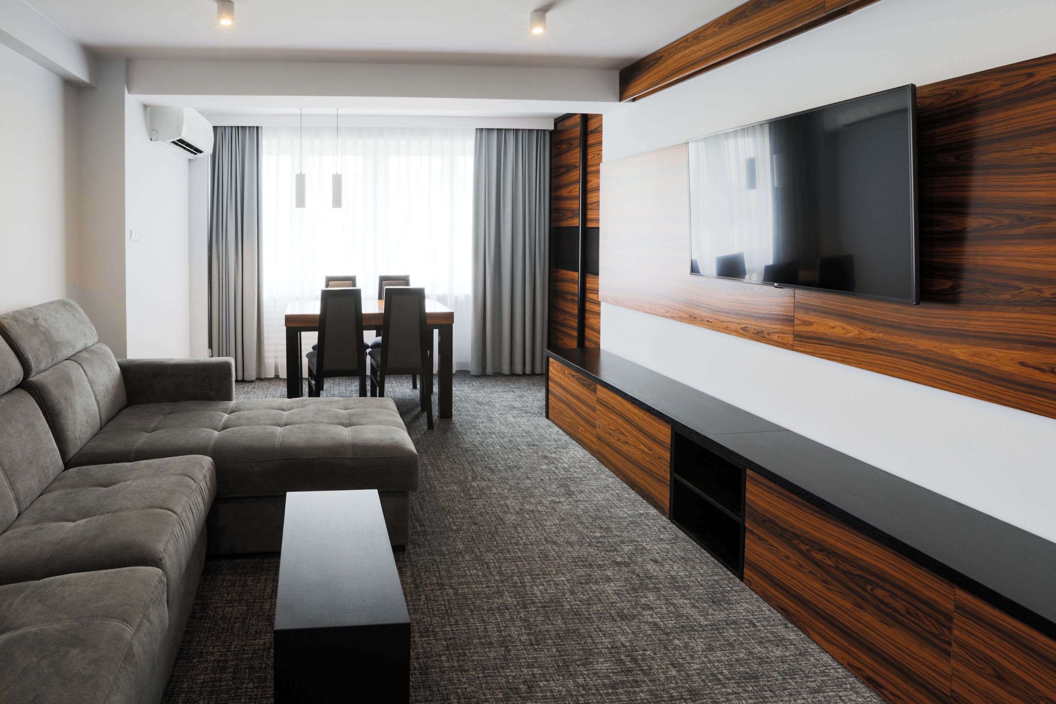 Ein modernes Hotelzimmer mit Ecksofa, einem flachbildfernseher an einer Holzvertäfelung, einem Essbereich mit Tisch und Stühlen sowie einem großen Fenster mit Vorhängen.